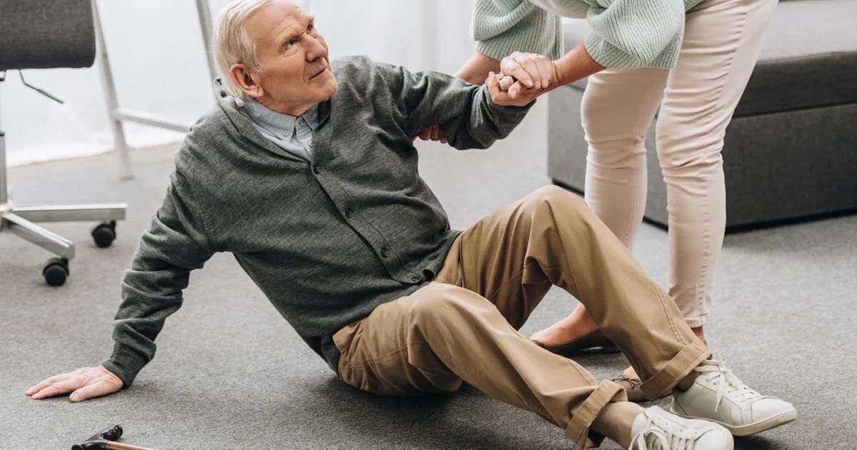 An older woman helping a senior man up from the floor after a fall, his walking cane lying beside them